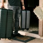 man in black denim jeans and black leather shoes standing beside black luggage bag